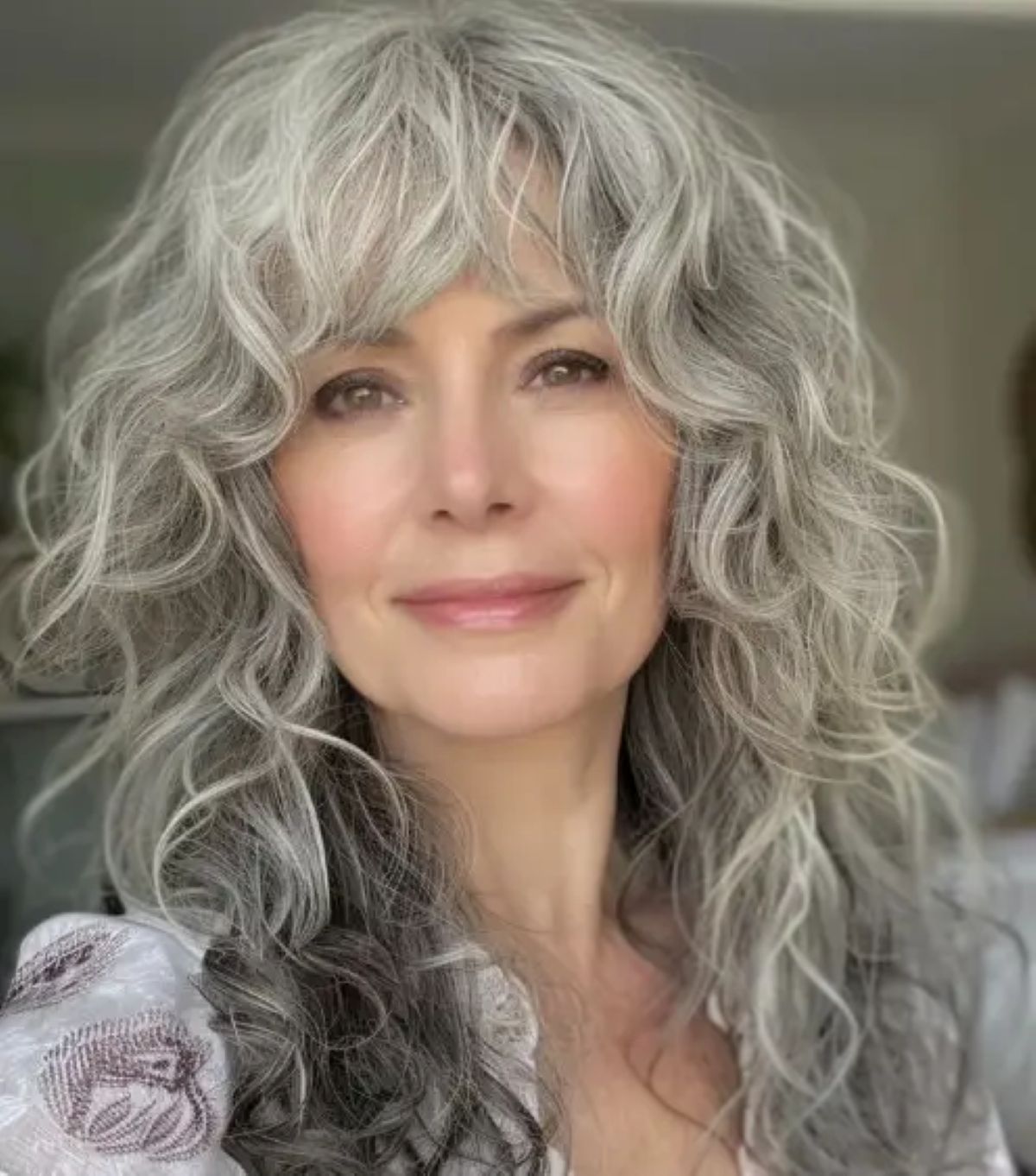 Mulher sorridente com cabelo grisalho, longo e ondulado, em corte Shaggy moderno com franja, transmitindo jovialidade.