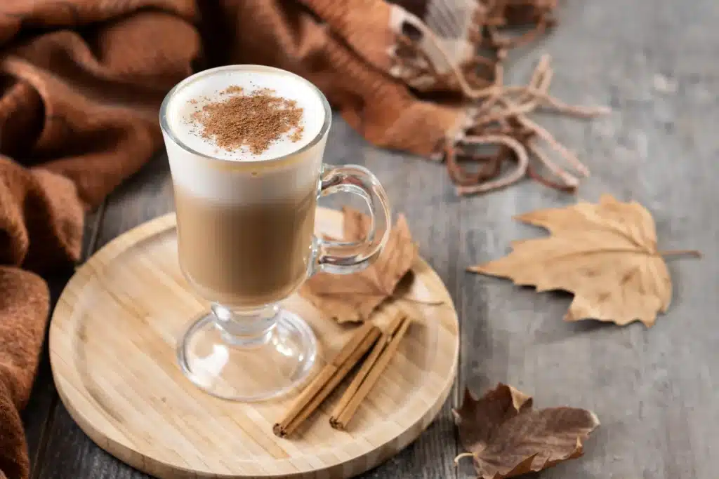 Xícara de café cremoso com canela sobre uma base de madeira, com folhas secas ao fundo.