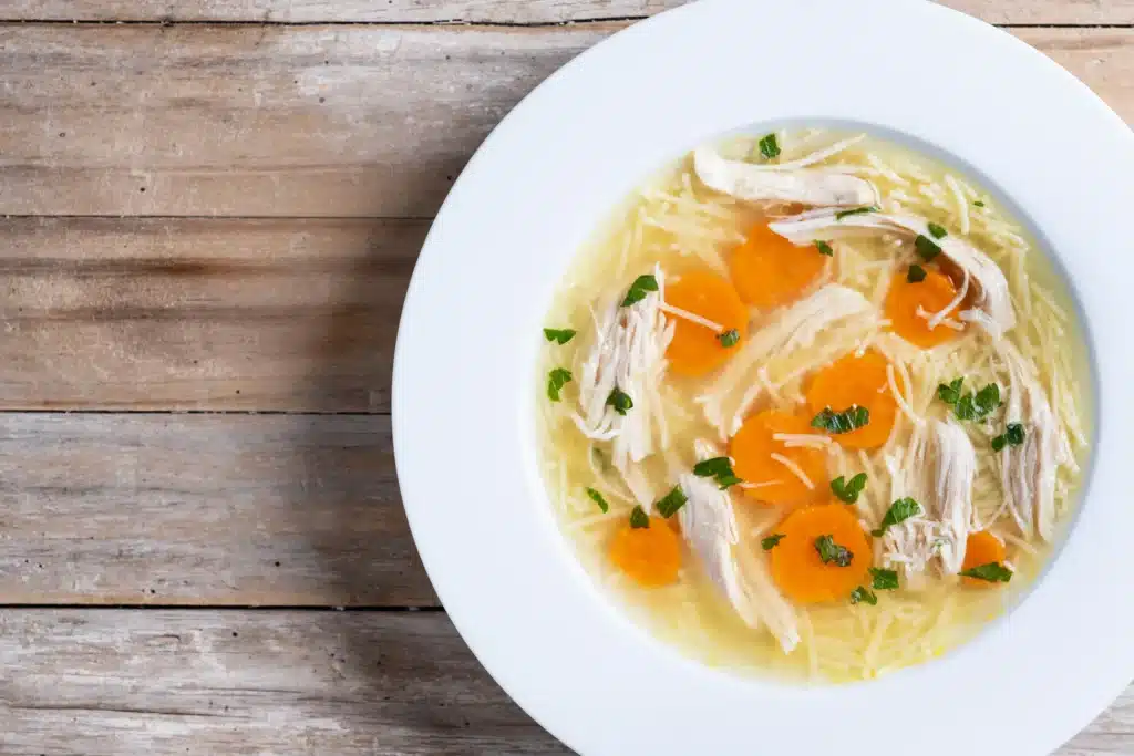 Canja de galinha com cenoura, frango desfiado e macarrão, com ervas frescas, em um prato branco.