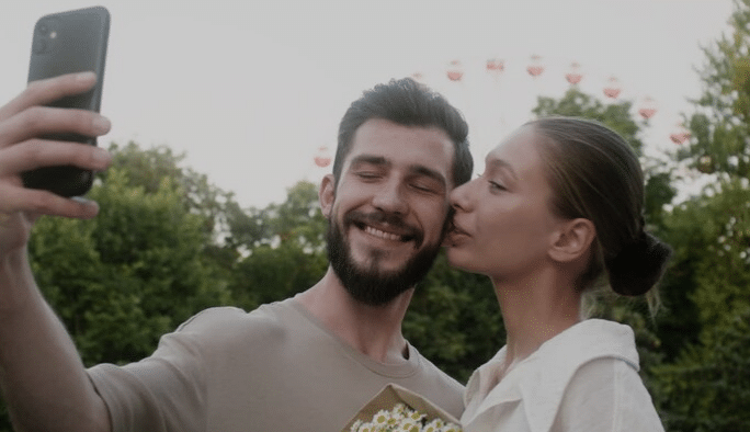 Jovem casal sorridente tirando selfie enquanto ela beija o rosto dele em parque com árvores ao fundo