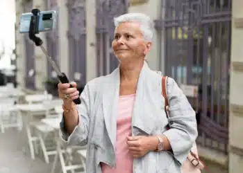 Mulher mais velha sorrindo enquanto usa um selfie stick para tirar uma foto em um ambiente urbano.