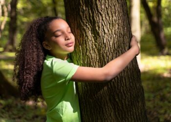 Menina de cabelos cacheados em uma camisa verde, abraçando o tronco áspero de uma árvore em uma floresta, com os olhos fechados, expressando calma e a sensação de alívio do estresse na natureza.