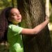 Menina de cabelos cacheados em uma camisa verde, abraçando o tronco áspero de uma árvore em uma floresta, com os olhos fechados, expressando calma e a sensação de alívio do estresse na natureza.