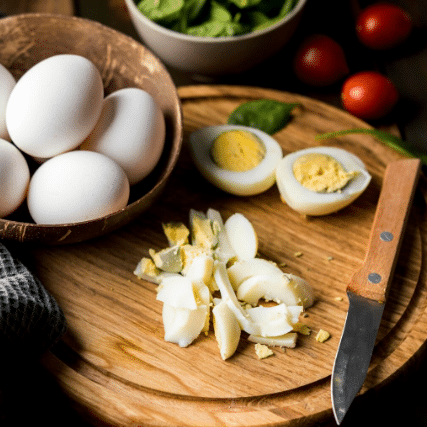 Ovos cozidos inteiros e picados sobre tábua de madeira ao lado de faca e tigela com mais ovos crus