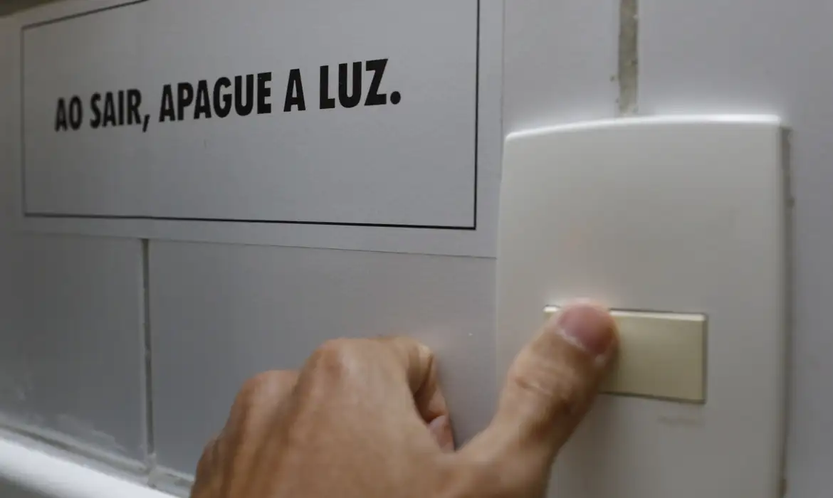 Mão apagando interruptor de luz próximo à placa com aviso sobre economia de energia