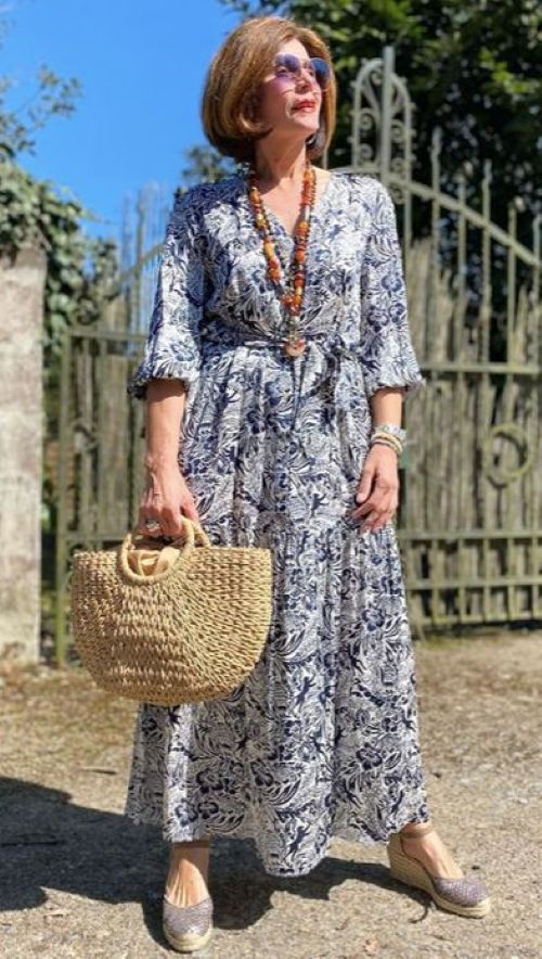 Mulher em um ambiente externo ensolarado usando um vestido longo de tecido fluido, estampado em tons de azul marinho e branco, com decote V e mangas três quartos. Acessórios incluem uma bolsa de palha e um colar longo.