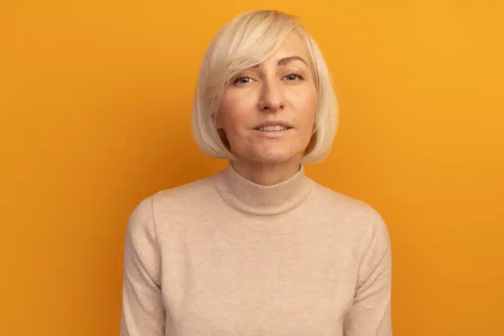 Mulher loira com franja lateral longa, sorrindo contra fundo amarelo.