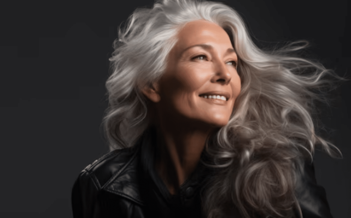Mulher madura sorridente com cabelos longs grisalhos esvoaçando e jaqueta de couro preta em fundo escuro