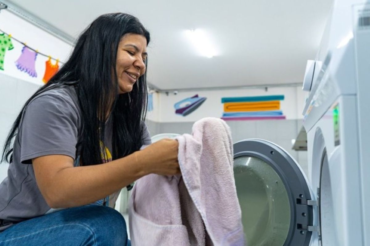 Mulher sorrindo, colocando roupas em máquina de lavar em lavanderia pública, simbolizando economia e tempo livre para mulheres.