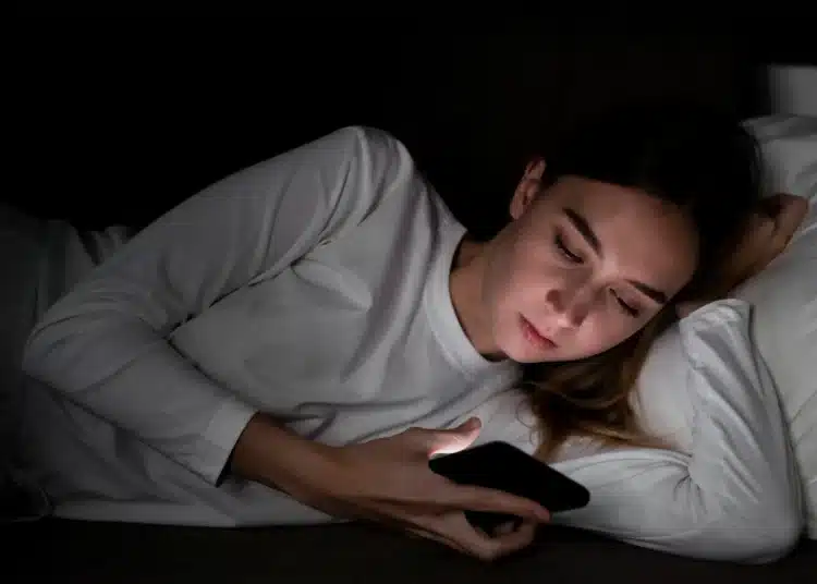 Mulher deitada na cama olhando para o celular no escuro.