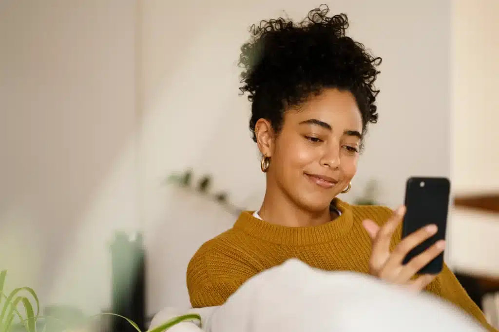 Mulher sorrindo enquanto usa smartphone, desejando boas vibrações para os amigos no WhatsApp.