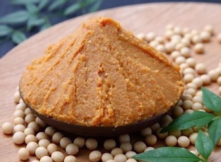 Monte de pasta de Missô em uma tigela sobre uma base de madeira, cercado por grãos de soja. O Missô é o ingrediente milenar fermentado de soja e koji.