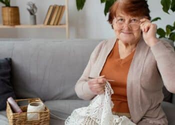 Senhora idosa sorridente, ajustando os óculos, fazendo crochê com linha branca e agulha na sala de casa, ilustrando um hobby da melhor idade.