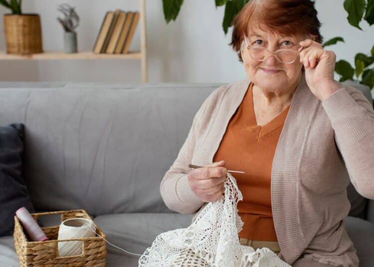 Senhora idosa sorridente, ajustando os óculos, fazendo crochê com linha branca e agulha na sala de casa, ilustrando um hobby da melhor idade.