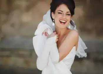Noiva sorrindo com vestido branco e acessórios elegantes, destacando-se com brincos grandes e luvas brancas.