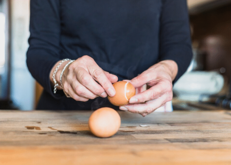 Mãos descascando um ovo cozido sobre mesa de madeira em ambiente de cozinha