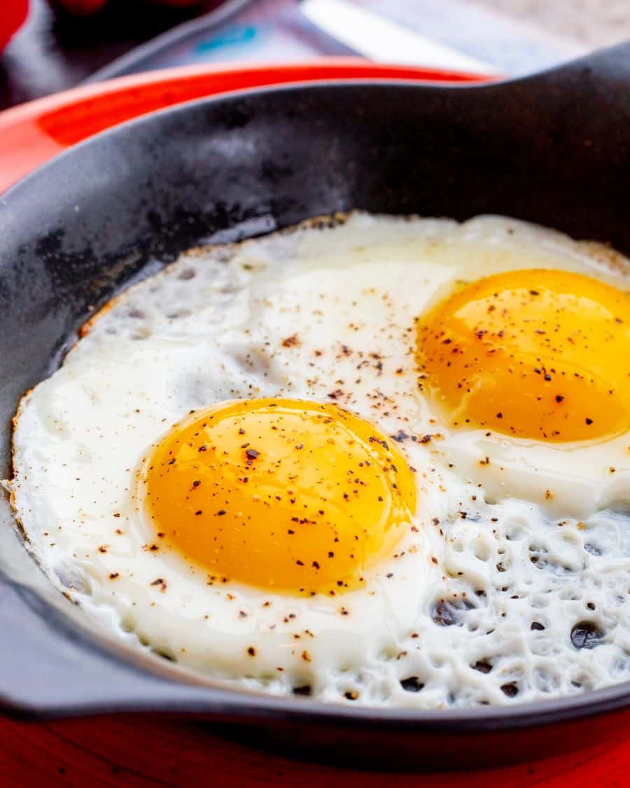 Dois ovos fritos perfeitamente em uma frigideira preta, salpicados com pimenta, com gema mole e clara firme. Ilustra o resultado de ovos que não grudam.