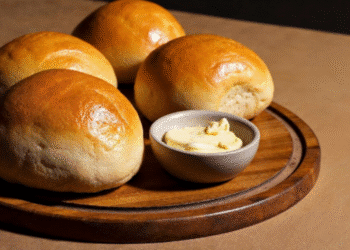 Quatro pães caseiros com casca dourada servidos em tábua de madeira ao lado de manteiga cremosa