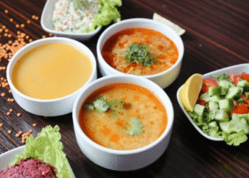 Três tigelas de sopas variadas e duas porções de saladas frescas sobre mesa escura de madeira