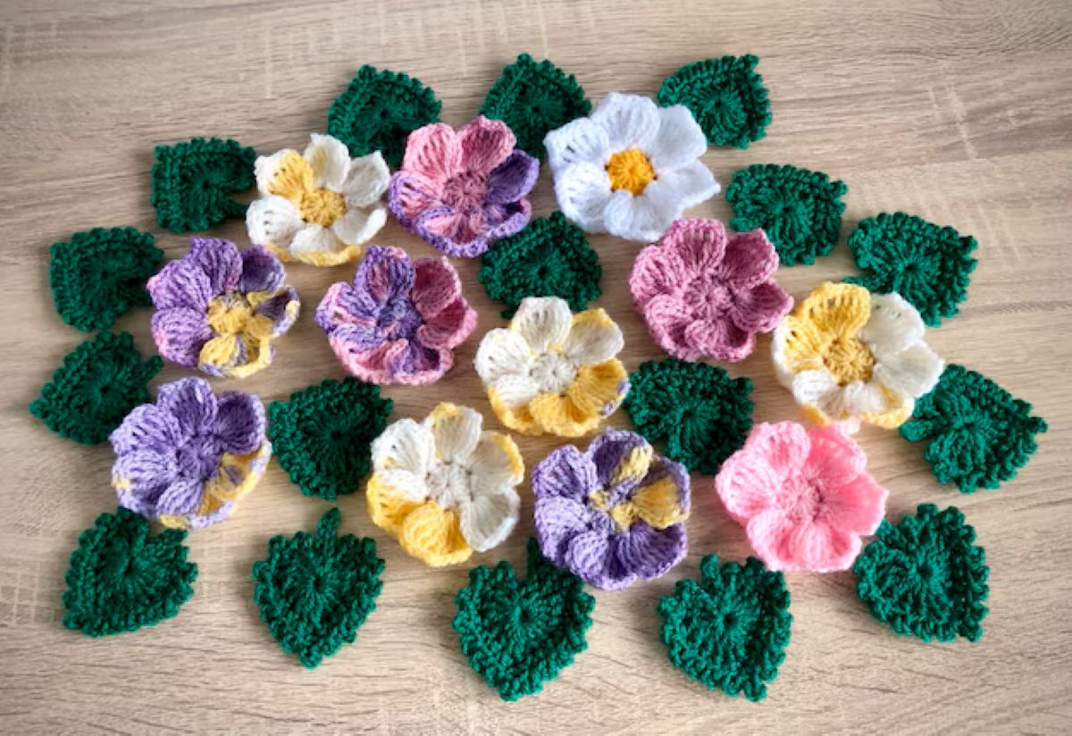 Arranjo de flores 3D de crochê e folhas em tons pastel (roxo, rosa, amarelo e branco) sobre superfície de madeira clara, ilustrando a aplicação em artesanato de moda.