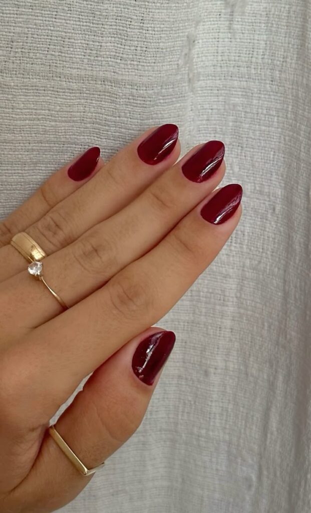 Mão com unhas curtas e arredondadas esmaltadas no tom vermelho cereja (vinho), destacando a cor que está em alta e o anel dourado.