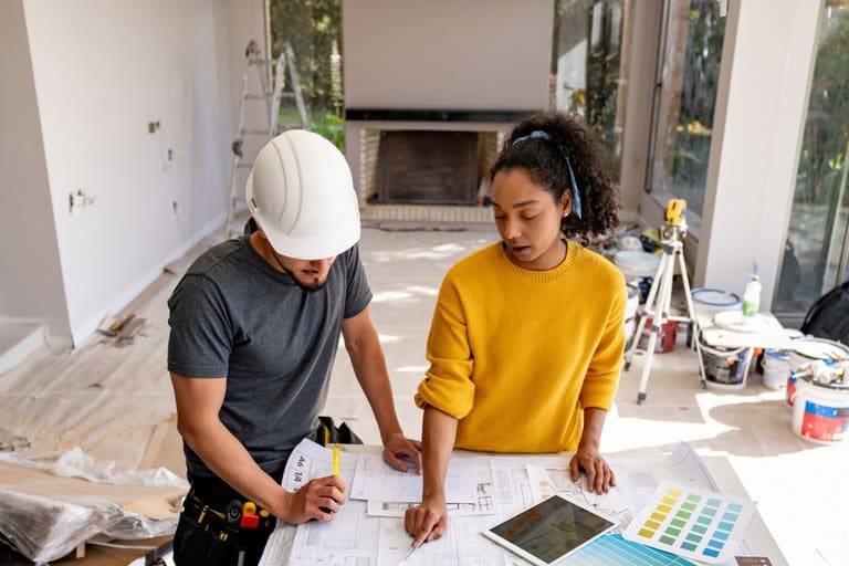 Casal ou proprietários revisando a planta de uma casa em reforma, com um profissional da construção civil ao lado, usando capacete. A imagem representa o planejamento de uma obra e a importância do financiamento facilitado pelo programa Reforma Casa Brasil.