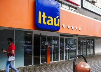 Agência bancária Itaú com fachada laranja e letreiro azul, homem de camisa vermelha andando na calçada.