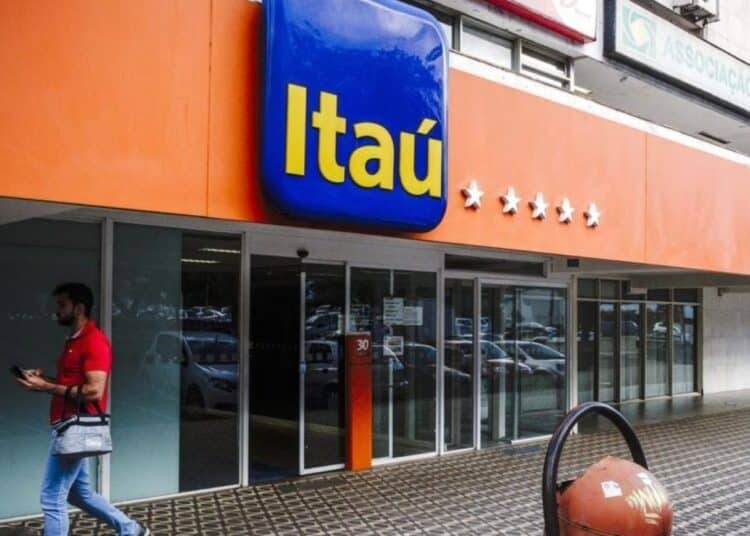 Agência bancária Itaú com fachada laranja e letreiro azul, homem de camisa vermelha andando na calçada.
