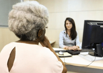 Aposentada idosa conversando com atendente em uma mesa de escritório, em frente a um computador, representando atendimento bancário ou financeiro.