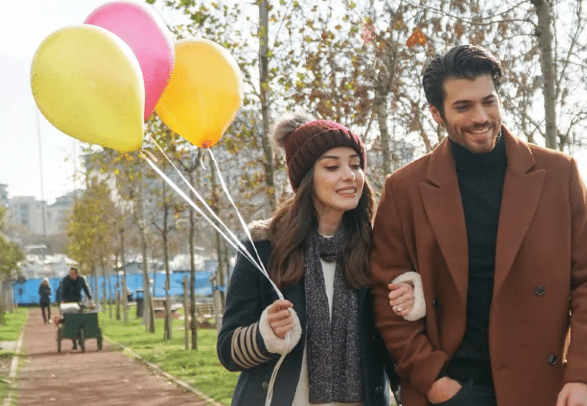 Um casal sorridente caminhando em um parque, a mulher segurando balões coloridos e o homem olhando para ela com carinho, representando a leveza e romance da novela turca 'Dolunay (Lua de Mel)'.