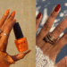 cDuas mãos exibindo esmaltes de cores vibrantes: uma com esmalte laranja intenso e outra com esmalte vermelho vinho escuro, indicando tendências para o fim de ano.