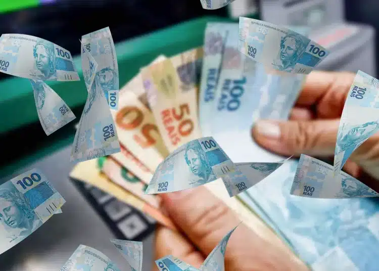 Notas de 100 reais e 50 reais flutuando ao redor de uma mão segurando dinheiro, com um caixa eletrônico ao fundo. Relacionado ao auxílio de R$ 250 aprovado no RS.