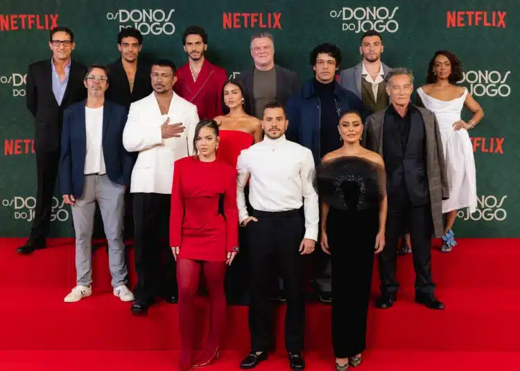 Elenco da série 'Os Donos do Jogo' posa para foto na estreia na Netflix, com trajes sofisticados, em evento de lançamento.