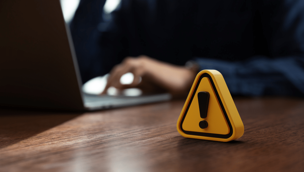 Símbolo de alerta amarelo sobre mesa de madeira escura, representando o risco constante de fraudes, enquanto uma pessoa ao fundo utiliza um notebook. Fraudes com cartao de credito exigem atenção.