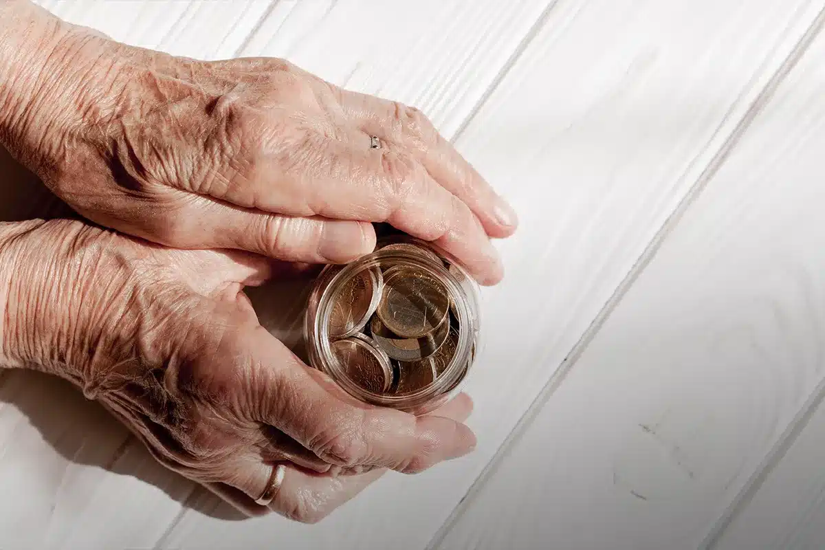 Mãos de idoso segurando um pote transparente com moedas de euro sobre mesa branca