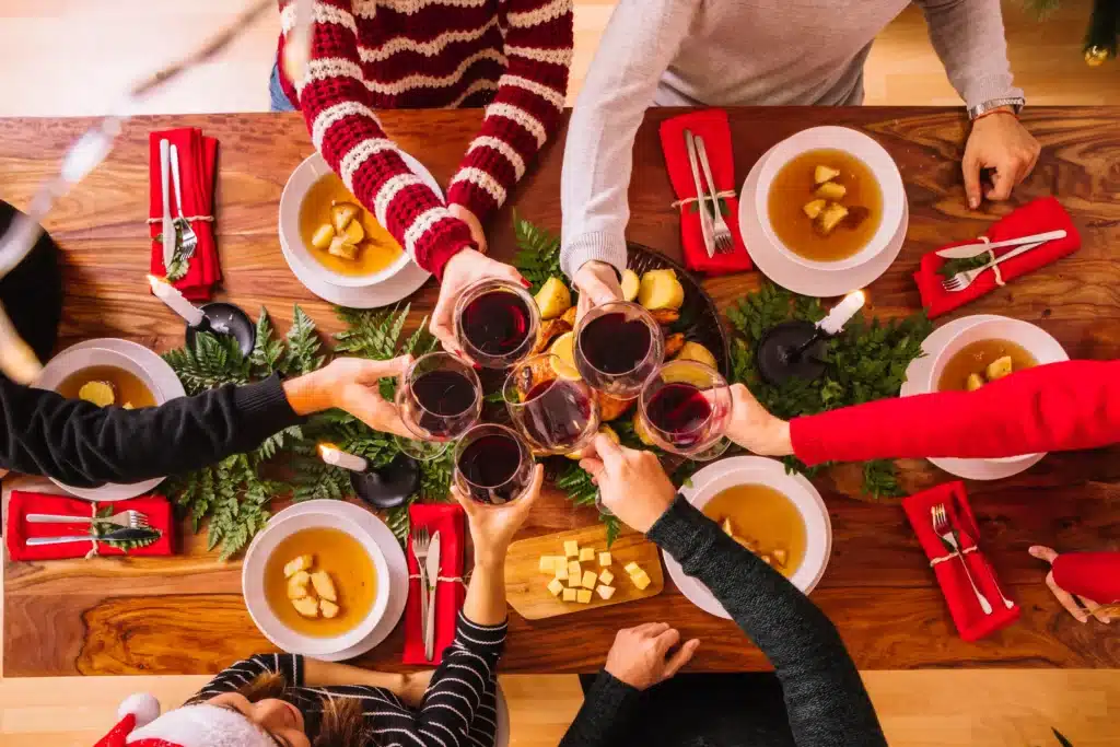 Grupo de pessoas brindando com taças de vinho durante uma ceia de Natal simples e econômica, com pratos tradicionais na mesa.
