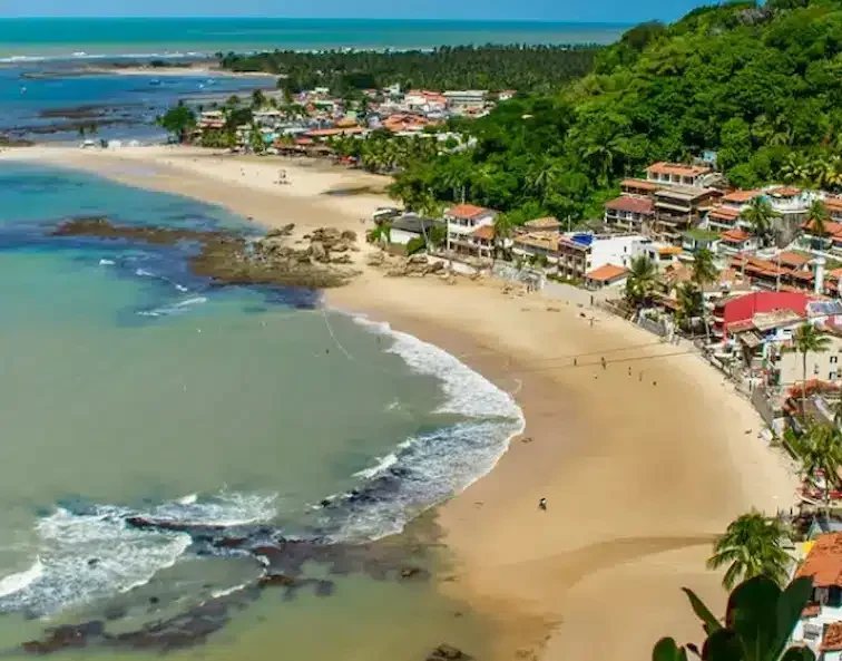 Vista aérea das praias e do vilarejo de Morro de São Paulo, com mar azul, faixa de areia ampla e casas entre a vegetação.