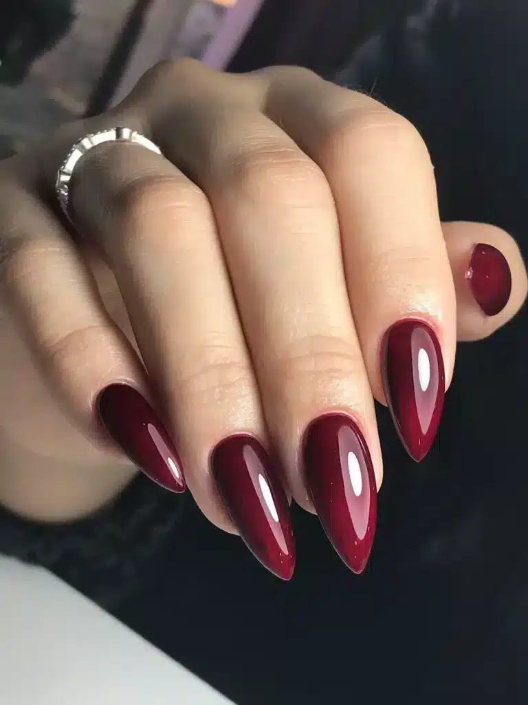 Vermelho vinho Unhas vermelhas em tom vinho com formato amendoado e anel prateado visível.