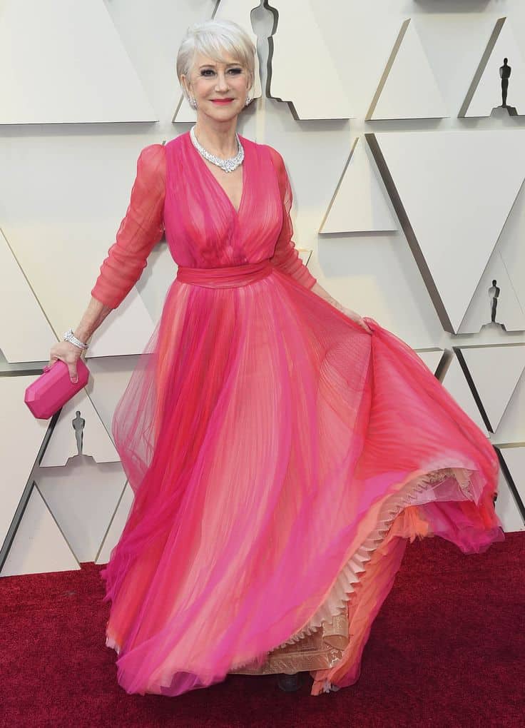 Mulher elegante com cabelo grisalho e vestido em tons de rosa vibrante, mostrando como o rosa confere leveza e calor ao visual, destacando a beleza dos fios brancos.