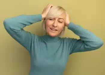 Mulher com cabelo grisalho iluminado e corte moderno, sorrindo e tocando os fios, com fundo verde claro.