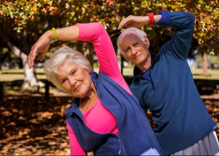 Casal de idosos sorrindo enquanto faz exercícios de alongamento ao ar livre, representando exercícios para pessoas acima de 60 anos.