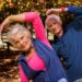 Casal de idosos sorrindo enquanto faz exercícios de alongamento ao ar livre, representando exercícios para pessoas acima de 60 anos.