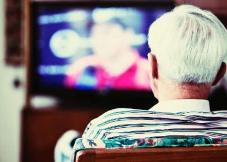 Pessoa idosa assistindo TV de costas, sentada em um sofá.