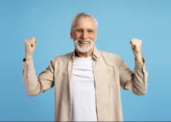 Idoso sorridente, com barba branca e óculos, celebrando com os punhos erguidos, em fundo azul.