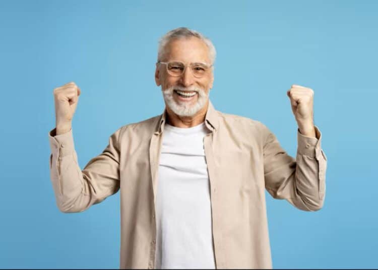 Idoso sorridente, com barba branca e óculos, celebrando com os punhos erguidos, em fundo azul.