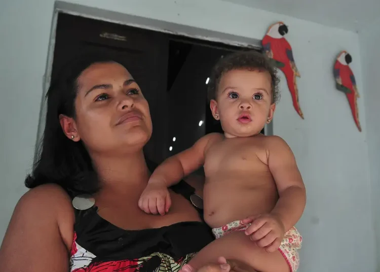 Mulher segurando bebê no colo em ambiente interno com papagaios vermelhos decorativos na parede azul