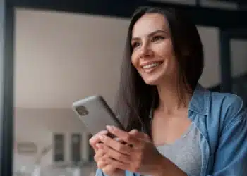 Mulher sorrindo enquanto segura o celular, simbolizando uma mensagem de alegria e otimismo para amigos.
