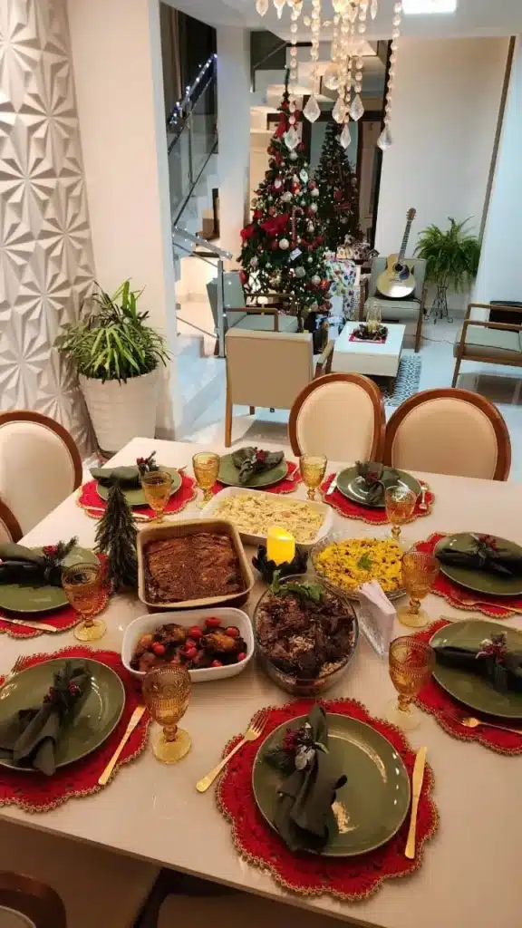 Mesa posta com crochê natalino, pratos verdes, taças douradas e comida festiva, com árvores de Natal ao fundo.
