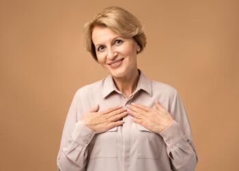 Mulher sorridente de cabelos loiros curtos, com mais de 50 anos, usando uma blusa em tom neutro e as mãos no peito, irradiando autoconfiança e a beleza da maturidade.