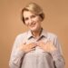 Mulher sorridente de cabelos loiros curtos, com mais de 50 anos, usando uma blusa em tom neutro e as mãos no peito, irradiando autoconfiança e a beleza da maturidade.